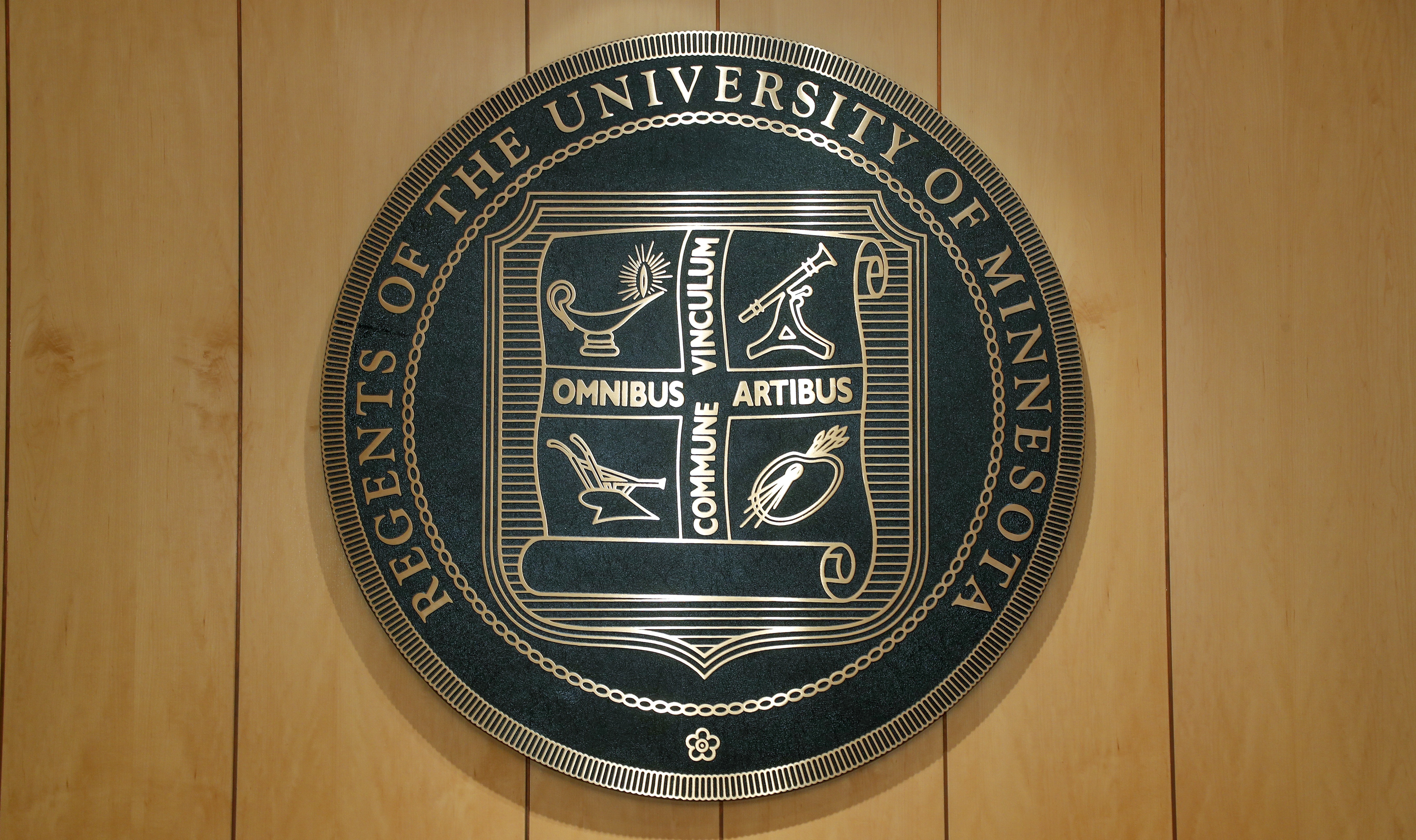 An image of a large, metallic regents seal hanging on a wood-panel wall.